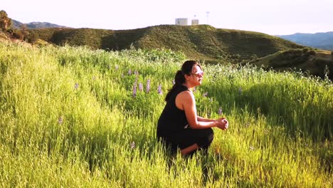 Slow-Motion-of-female-in-Black-Dress-and-Sunglasses-kneeling-in-Green-Field-and-Watching-Sunset-in-Distance