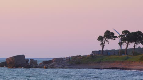 Entfernte-Menschen-Genießen-Romantischen-Sonnenuntergang-Am-Meer-Mit-Einer-Windkraftanlage-Im-Hintergrund,-Weitwinkelaufnahme