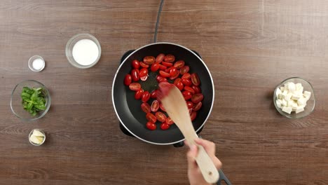 Top-down-shot-of-cherry-tomatoes-sauteed-in-a-pan-on-a-table-top-hob-on-wooden-table-with-other-ingredients-around,-adding-in-tomato-paste-and-stirring