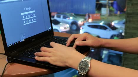 Male-person-surfing-the-net-and-performing-online-work-on-his-laptop-computer-at-a-coffee-shop,-with-heavy-vehicular-traffic-showing-through-a-glass-wall