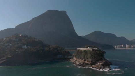 Rotational-aerial-pan-showing-the-Joatinga-beach-on-a-hazy-backlit-day-with-the-Gavea-mountain-behind-revealing-the-wider-well-known-cityscape-of-Rio-de-Janeiro-in-the-background