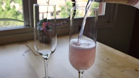 Two-glasses-of-pink-champagne-being-poured-on-a-kitchen-table-by-the-window