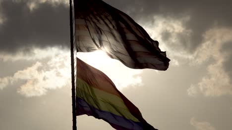 Regenbogen-LGBT-Gay-Pride-Flagge-Weht-Im-Wind-4-Und-Die-Amerikanische-Flagge-Weht-Mit-Sonne-Hinterleuchtet-Unter-Grauen-Wolken-4k