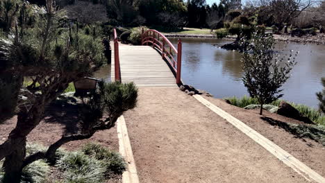 Pan-shot,-walkway-to-red-bridge-over-pond,-Ju-Raku-En-Japanese-Garden,-Toowoomba-Australia