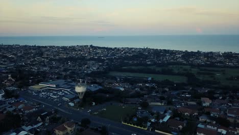 Aerial-footage-from-a-drone-of-residential-homes-with-a-road-with-moving-traffic-overlooking-a-water-tower-shopping-centre-and-a-golf-course-with-a-green-grassy-park