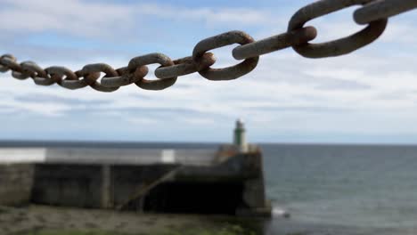 Chain-Link-In-Focus-With-Waves-Hitting-Stone-Pier-With-Small-Lighthouse-At-Laxey,-Isle-Of-Man