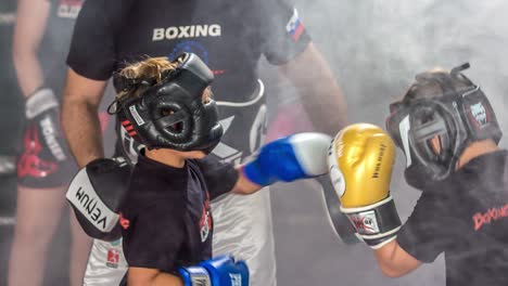 Two-kids-doing-kickbox-sparing-training,-punching-each-other-in-the-head,-wearing-protective-gear---slow-motion-video,-camera-following-action,-smokes-all-around-them