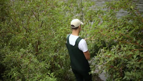 Slow-Motion-Shot-of-a-male-fisherman-wearing-waders-walking-through-the-bushes-down-to-the-river-to-do-some-Fly-Fishing