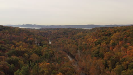 aerial-dolly-in,-on-a-cloudy-day,-drone-camera-tilts-down-over-the-stream---tree-tops-with-orange-leaves-at-The-New-Croton-Dam-in-Westchester-County,-NY