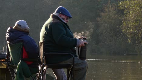 Retired-pensioner-gentleman-sitting-in-autumn-park-drinking-warm-steaming-beverage-from-flask-looking-out-across-park-lake