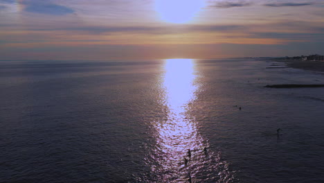 high-up-dolly-shot-forward,-descend-over-surfers-in-the-Atlantic-Ocean,-drone-camera-moving-towards-a-golden-sunset