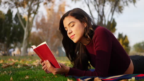 Eine-Hispanische-College-Studentin,-Die-Ein-Rotes-Buch-Liest-Und-Lächelt,-Während-Sie-Sich-Bei-Sonnenuntergang-Im-Gras-Eines-Parks-Niederlegt