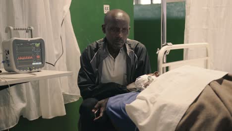Father-sitting-next-to-his-son-on-a-hospital-bed-in-a-clinic-after-eye-surgery-with-a-heart-monitor-next-to-them