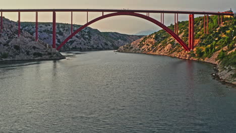 wide-aerial-shot-of-a-big-red-bridge-on-a-beautiful-mountain-scenery-on-sunset,-camera-moving-forward-panning-up