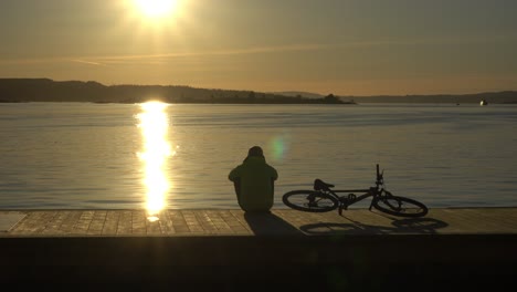 Male-silhouette-sitting-beside-bike-on-harbour,-watching-sunset-over-Oslo-Fjord
