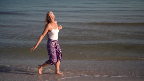 Slow-motion-of-mature-woman-in-sarong-walking-on-a-beach-and-splashing-in-the-water-at-sunrise-or-sunset