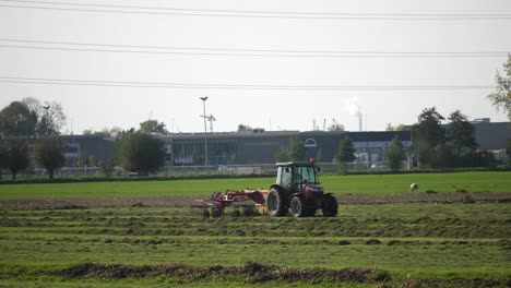 Un-Tractor-Rojo-Está-Conduciendo-Y-Recolectando-Cultivos-En-El-Campo-Verde-Con-Ovejas-En-El-Fondo-Y-Cables-De-Alimentación-Arriba