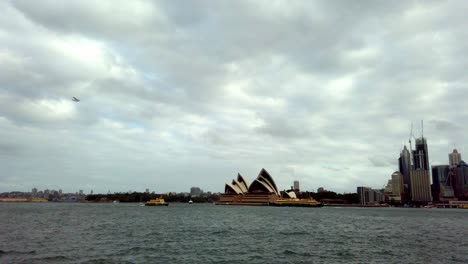 Opera-House-and-downtown-waterfront-view-with-a-seaplane-passing-by-on-an-overcast-day,-handheld-shot-from-a-vessel