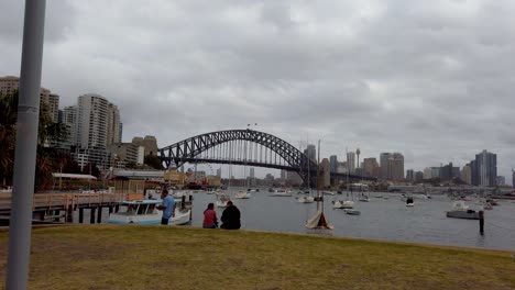 Downtown-waterfront-view-with-Harbour-Bridge-and-people-enjoying-the-view-on-an-overcast-day,-Shot-from-a-moving-vehicle