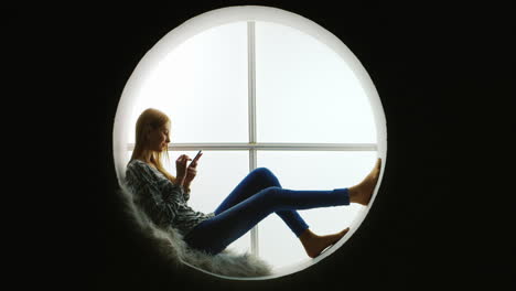 Girl-Sitting-in-Round-Window-Using-Smartphone