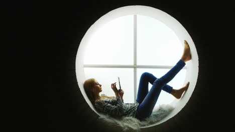 Woman-Relaxing-in-a-Round-Window-Uses-Tablet