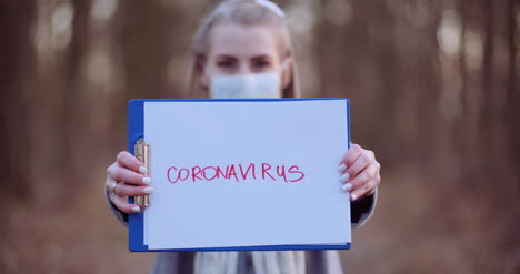 Woman-With-Protective-Mask-Holding-Coronavirus-Inscription-In-Hands-In-Forest