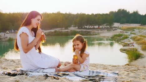 Mum-With-A-Daughter-Three-Years-Relaxing-On-The-Beach-By-The-Lake-Drink-Juice-From-Bottles-With-Tube