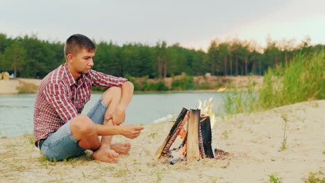 Young-Attractive-Man-Sitting-By-The-Fire-Alone-Fries-Marshmallow-On-A-Stick-Against-The-Background-O