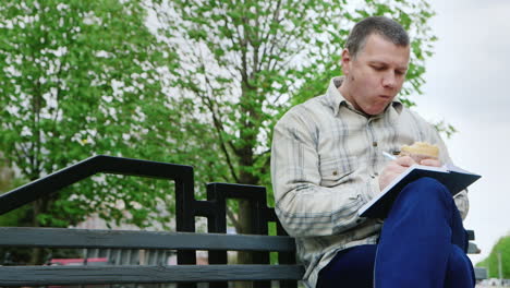 Man-Eating-Fast-Food-On-A-Park-Bench-Writes-Something-In-A-Notebook