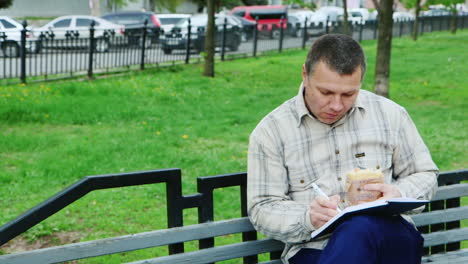 Man-Eating-Fast-Food-On-A-Park-Bench-Writes-Something-In-A-Notebook