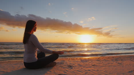 Junge-Frau-Mit-Langen-Haaren,-Die-Bei-Sonnenuntergang-Am-Strand-Sitzt-Und-Ruhiges-Selbstvertrauen-Und-Gesundheit-Meditiert