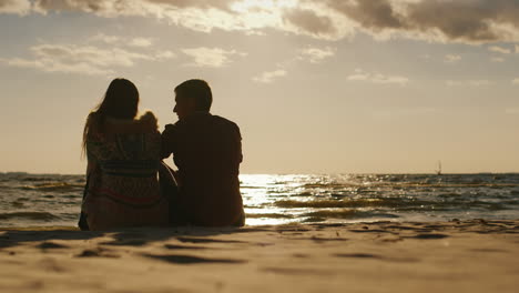 Silhouetten-Von-Verliebten-Paaren,-Die-Am-Strand-Sitzen-Und-Den-Sonnenuntergang-Beobachten,-Und-Ein-Brett-Mit-Segelrücken