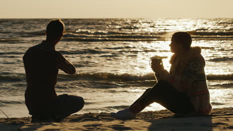 Romantisches-Junges-Paar,-Das-Sich-An-Einem-Kühlen-Tag-Am-Strand-Oder-Einem-See-Entspannt,-Frau-Trinkt-Tee-Aus-Einer-Tasse-A