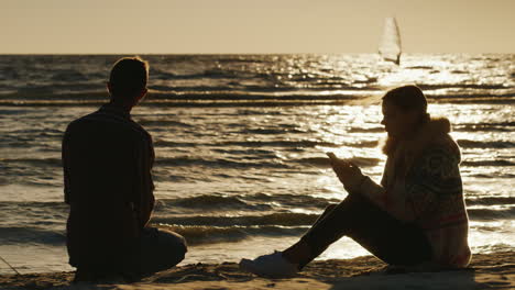Romantic-Couple-Relaxing-On-The-Beach-At-Sunset-Woman-Enjoying-A-Telephone-A-Man-Throws-Stones-Into-