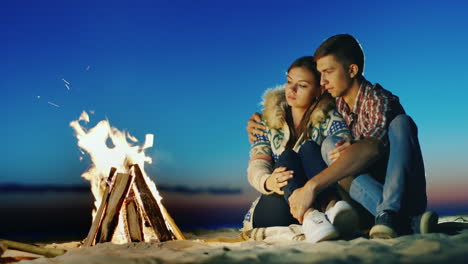 Romantic-Young-Couple-Sitting-On-The-Beach-Campfire-Hug-Admiring-The-Fire-Dreaming