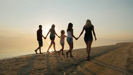 The-Company-Of-Children-And-Adults-Are-Having-Fun-On-The-Beach-Holding-Hands-Rear-View-At-Sunset