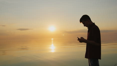 A-Teenager-Of-15-Years-Enjoys-The-Phone-It-Should-At-Sunset-On-The-Background-Of-A-Calm-Sea