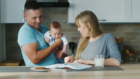 Young-Family-In-The-Kitchen-Mom-Reading-A-Book-With-The-Baby-Daddy-Is-Near
