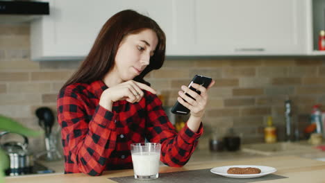 A-Young-Woman-Uses-A-Smartphone-He-Is-Sitting-In-His-Kitchen-Standing-Next-To-A-Glass-Of-Milk-Health