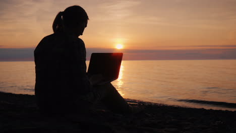Silhouette-Of-A-Woman-Working-With-A-Laptop-By-The-Sea-At-Sunset
