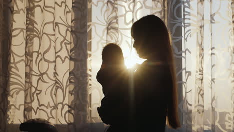 Silhouette-Mother-With-Baby-Standing-At-The-Window-At-Sunset-The-Sun's-Rays-Shine-Beautifully