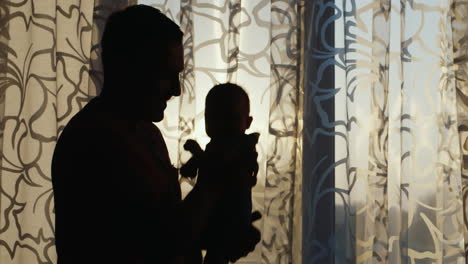 Silhouette-Of-A-Healthy-Young-Father-Playing-With-Baby-By-The-Window-At-Sunset-Happy-Childhood