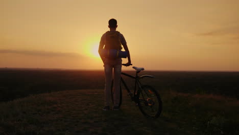 Ein-Mann-Mit-Rucksack-Und-Fahrrad-Steht-Und-Schaut-Auf-Den-Horizont,-Wo-Die-Sonne-Untergeht,-Radfahren-Und