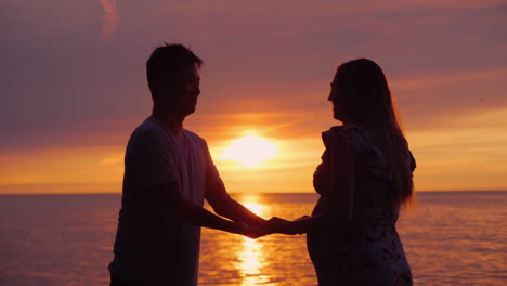 Side-View-Of-An-Asian-Man-Holding-His-Pregnant-Wife's-Hands-In-The-Background-Of-Sunset-Over-The-Sea