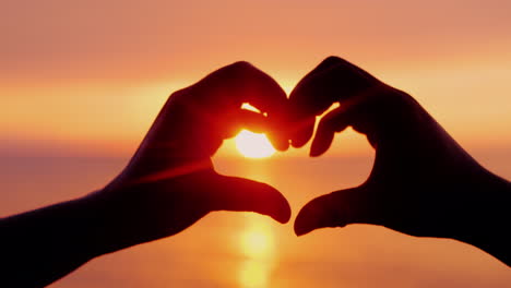 Two-Hands-Show-The-Shape-Of-The-Heart-Around-The-Sun-Setting-Over-The-Sea
