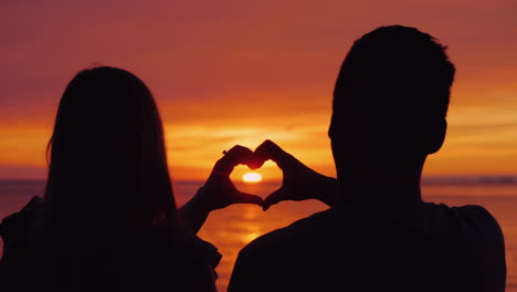 Silhouettes-Of-A-Young-Couple-Hands-Show-The-Shape-Of-The-Heart-Against-The-Backdrop-Of-The-Sunset-O