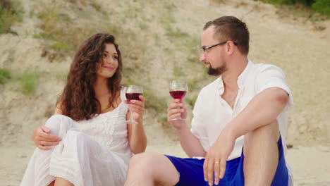 A-Young-Couple-Is-Drinking-Wine-By-The-Sea-At-Sunset-They-Sit-On-The-Sand-Clink-Glasses-Anniversary-
