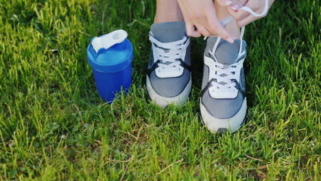 Tie-Shoelaces-On-Sports-Shoes-Female-Legs-On-A-Green-Lawn-Near-A-Bottle-For-Water-Ready-For-An-Activ