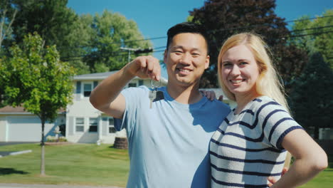 Happy-Multi-Ethnic-Couple-On-The-Background-Of-Their-New-Home-Holds-A-Key-Chain-With-Keys-In-Hand-Bu