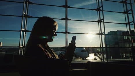 The-Silhouette-Of-A-Woman-Sitting-Facing-A-Large-Window-In-The-Airport-Terminal-Uses-A-Smartphone-Wa
